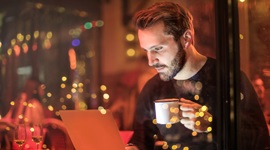 A photo of a man inside a cafe holding a cup of coffee while looking at something on his laptop